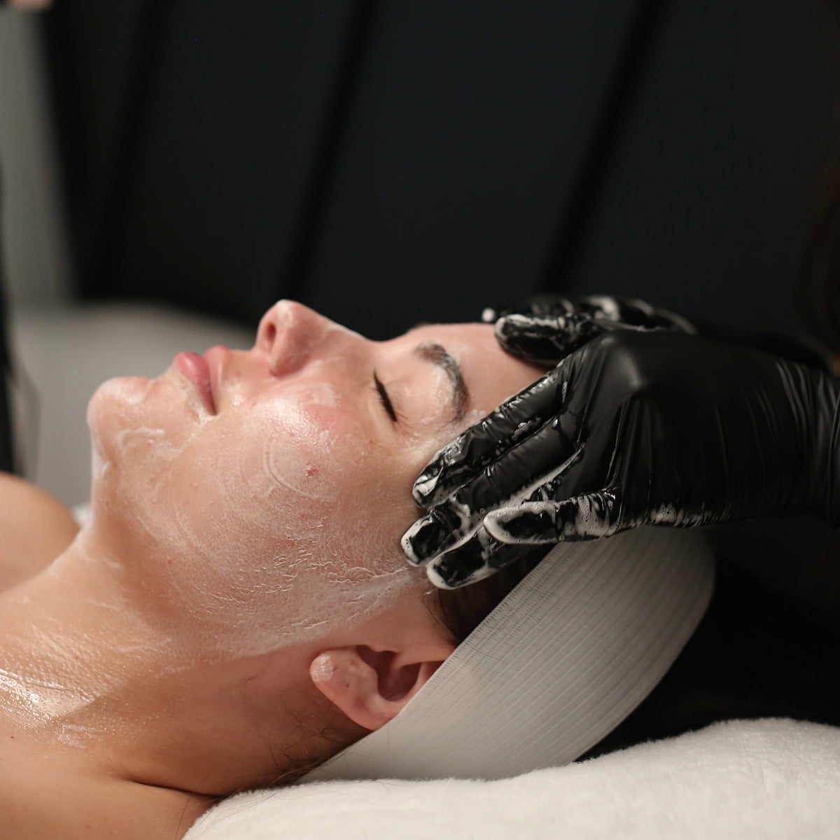A close-up of a person lying down with a white face mask on their face, while a person wearing black gloves gently cleans their forehead. The background is a dark, modern interior with black paneling.